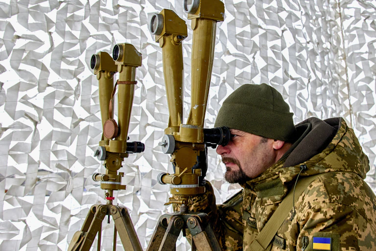 A man wearing a Ukranian military uniform looks through a specialised long-distance viewing apparatus during a training exercise.