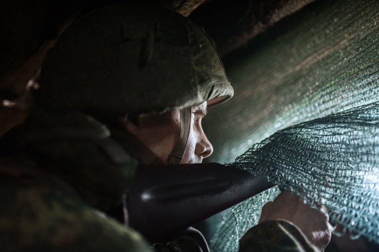 Man wearing army helmet pointing gun out through a hole.
