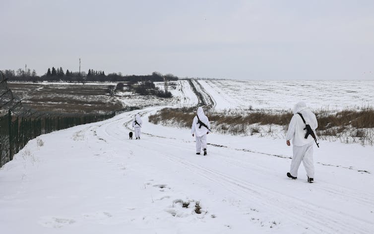 Three armed military officials walk in the snowy countryside wearing white camouflage.
