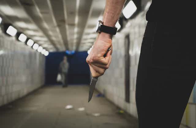 Close-up of a man's hand holding a knife while walking through a footpath tunnel. Another person can be seen in the distance.