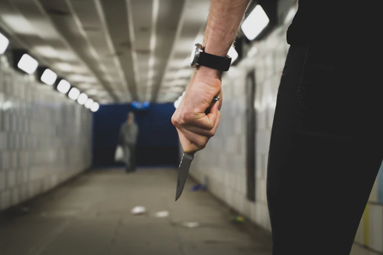 Close-up of a man's hand holding a knife while walking through a footpath tunnel. Another person can be seen in the distance.