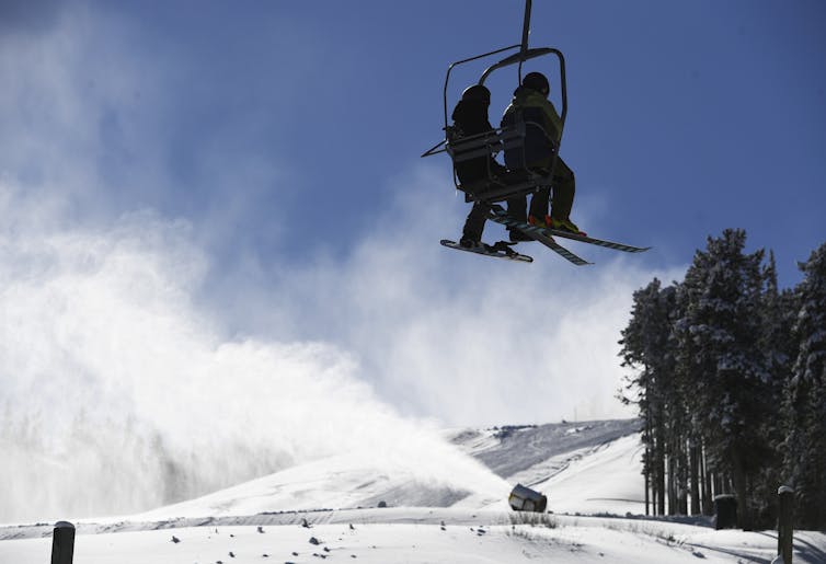 Skiers on a lift with a snowmaking machine running below