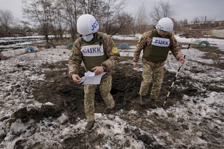 two men in military uniforms, protective vests and white-sheathed helmets step out of a small crater in snow-covered ground