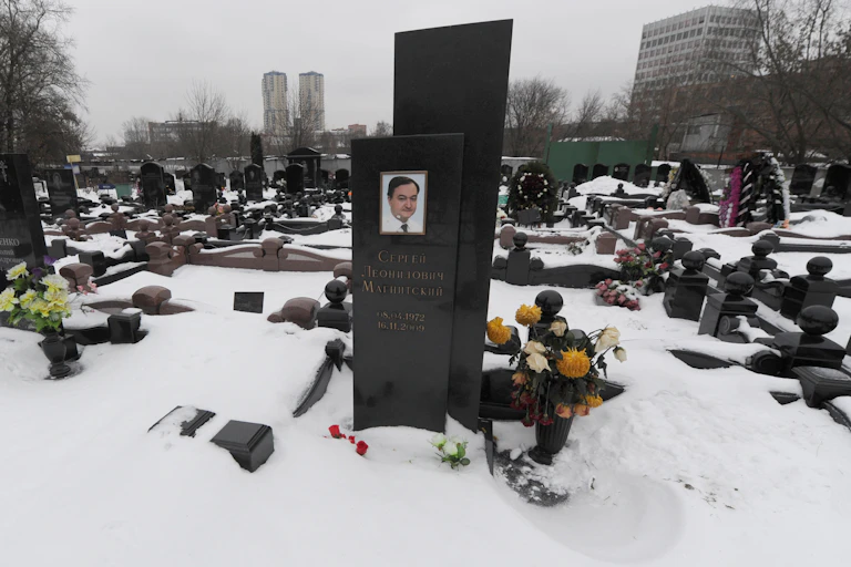 Flowers are placed by an obelisk memorial bearing a photo of Sergei Magnitsky in a snowy cemetery.