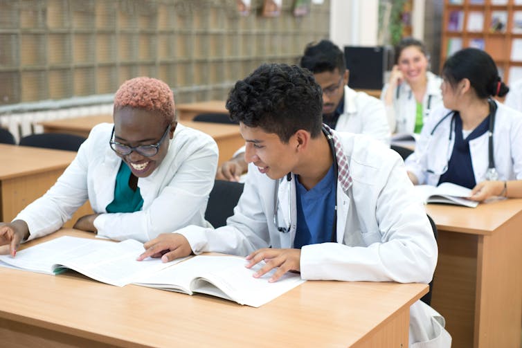 A Black and South Asian student sit in lab coats poring over medical books.