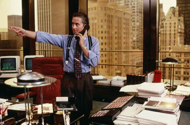 actor Michael Douglas looks to the right and points with his left hand and a cigarette in his fingers while holding a phone with his left hand while standing in his office