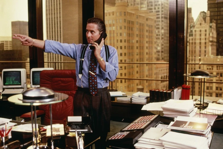 actor Michael Douglas looks to the right and points with his left hand and a cigarette in his fingers while holding a phone with his left hand while standing in his office