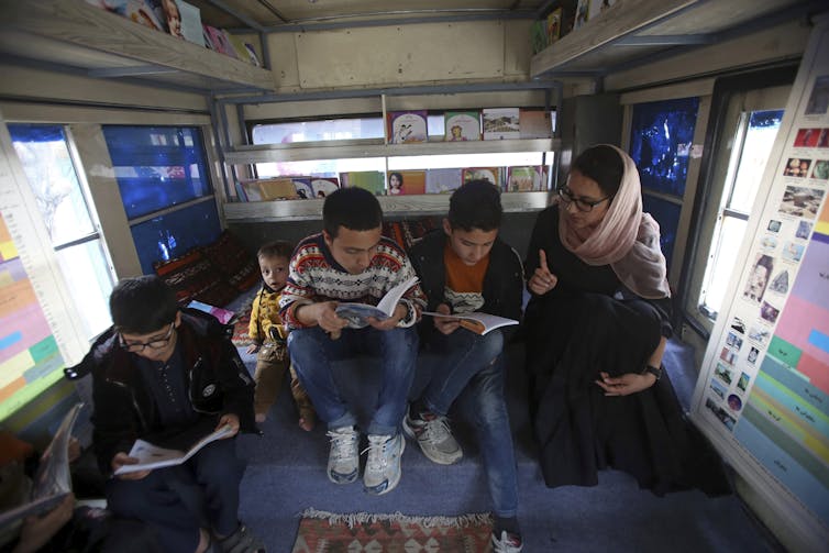 A woman and children seen sitting on a bench in the inside of a refurbished bus with books.