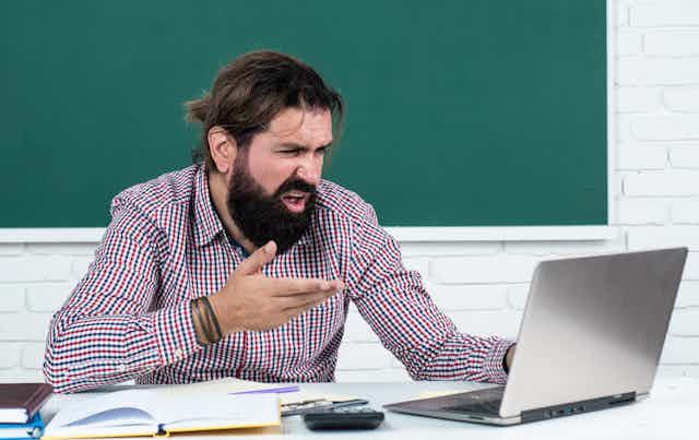 Bearded man gestures in dismay as he reads from his laptop