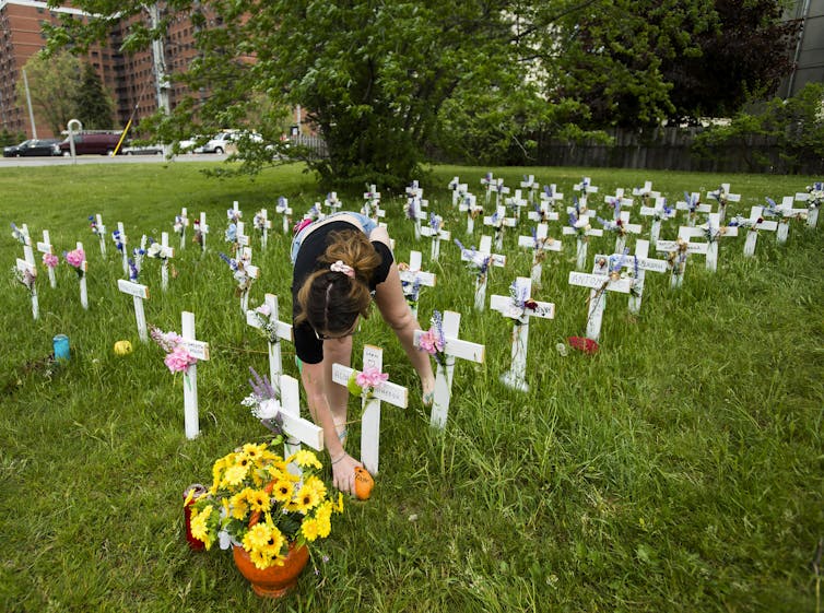 A young woman bent over one of dozens of small white crosses on a green lawn