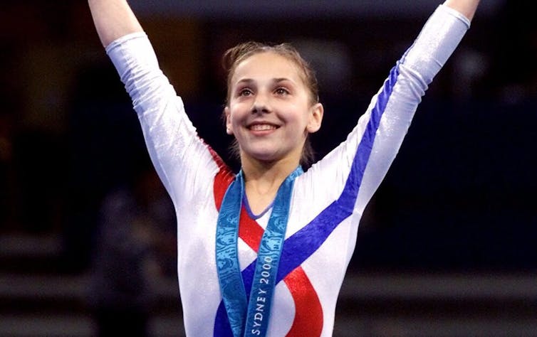 A girl in a gymnastics leotard with her arms raised. She has a gold medal around her neck and she is smiling.