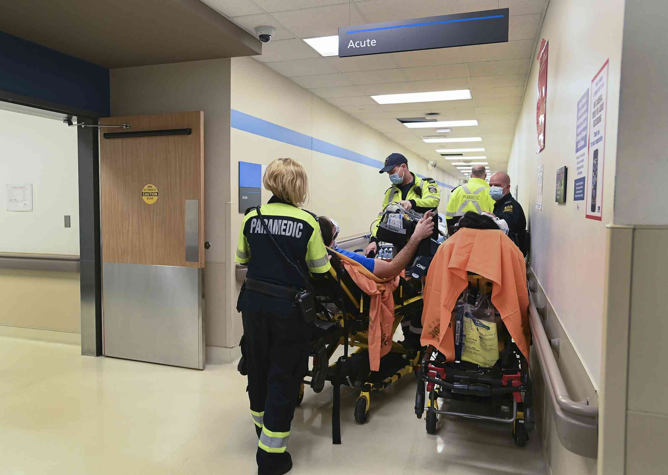 Paramedics wheeling a patient on a gurney draped in orange in a crowded hospital corridor