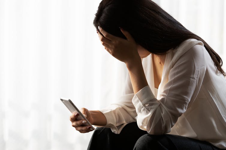 A woman with long black hair holds a phone in one hand and her other hand cover her eyes.