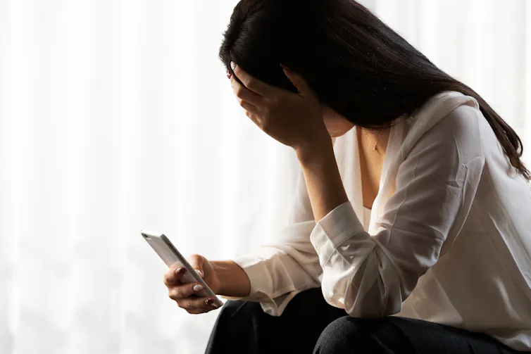 A woman with long black hair holds a phone in one hand and her other hand cover her eyes.