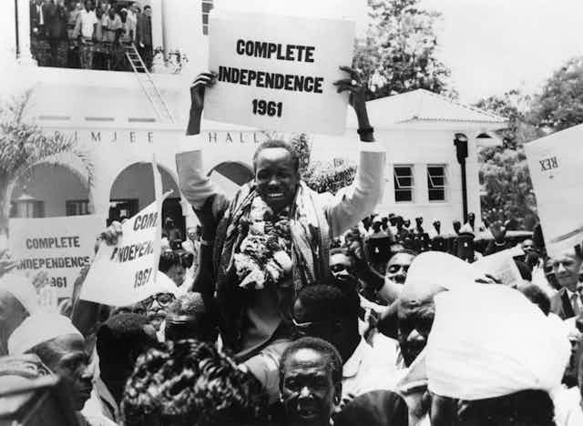 A black and white archive photo of a man holding a placard that reads 'Complete independence 1961' and beaming. He is in the street, being held aloft by a throng of people as others watch from a balcony.