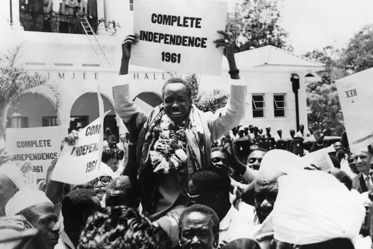 A black and white archive photo of a man holding a placard that reads 'Complete independence 1961' and beaming. He is in the street, being held aloft by a throng of people as others watch from a balcony.