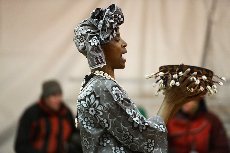 A woman sings and dances, dressed in traditional East African fabric with headpiece and holding a wooden bowl, the sides strung with cowrie shells.