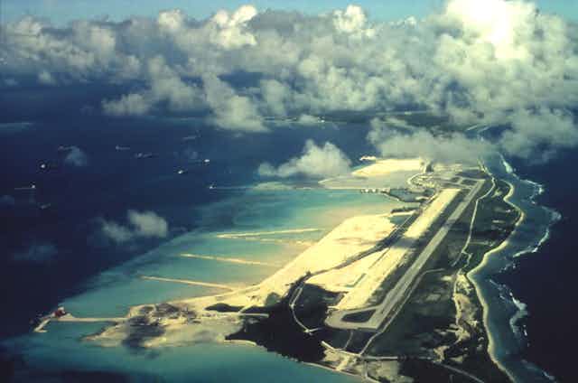 An overhead view of a runway on a slim island surrounded by deep blue ocean.