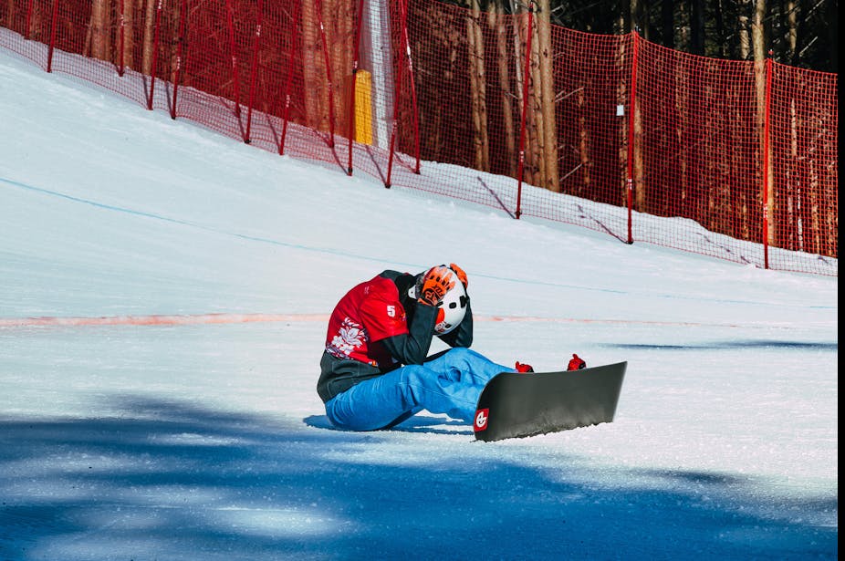 A snowboarder sits on the ground after finishing their run, head down and holding their helmet.  