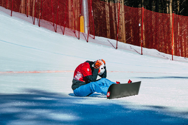 A snowboarder sits on the ground after finishing their run, head down and holding their helmet.