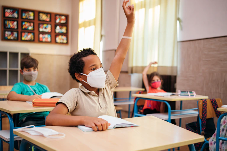 Three mask-wearing students, one with his hand raised, sit at their desks in a classroom.