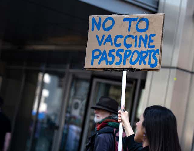 A woman holds a cardboard sign with NO TO VACCINE PASSPORTS written on it in blue paint