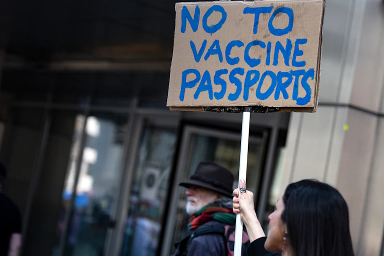 A woman holds a cardboard sign with NO TO VACCINE PASSPORTS written on it in blue paint