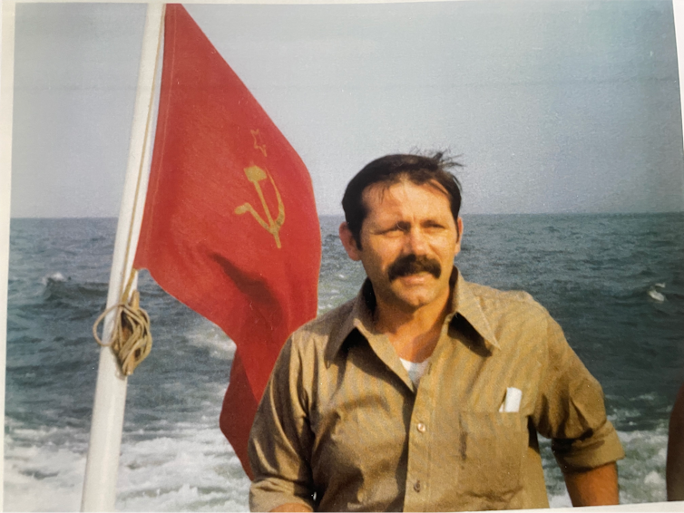 A man with a mustache is pictured next to a Soviet flag, in front of the ocean.