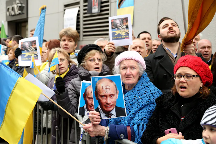 Women and men hold Ukrainian flags and photos of Vladimir Putin with a swastika on his forehead.