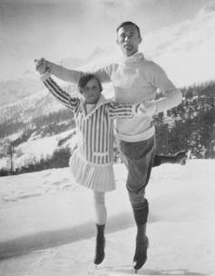 Two skaters on ice outside with mountains in the background. They are posing as if gliding together.