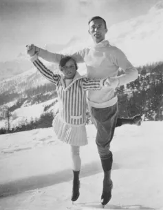 Two skaters on ice outside with mountains in the background. They are posing as if gliding together.