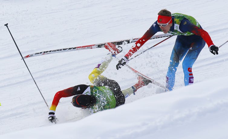 A cross-country skier falls in front of another during a race. The second skier has his mouth open as if shouting.