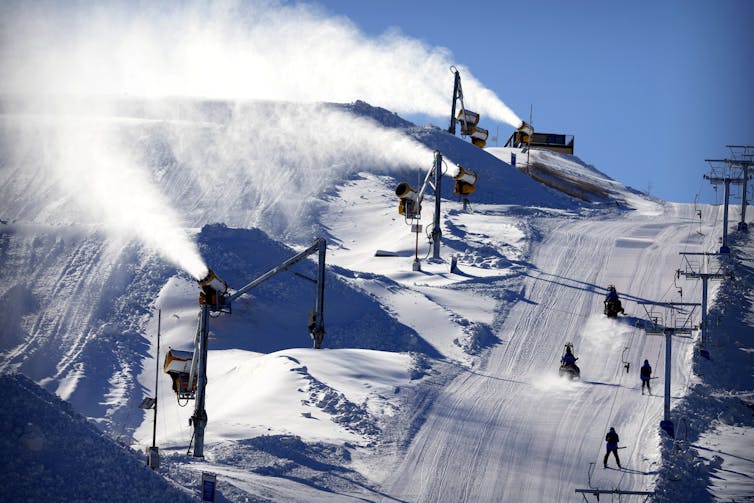 Several barrels blow snow onto one ski run while skiers uses another.