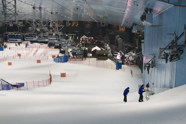 Two workers pack snow on an indoor ski slope with a sloped ceiling overhead.