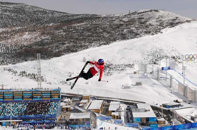 A freestyle skier crosses her skis while high in the air, with mountains and a stadium below as the backdrop.