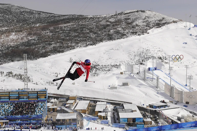 A freestyle skier crosses her skis while high in the air, with mountains and a stadium below as the backdrop.