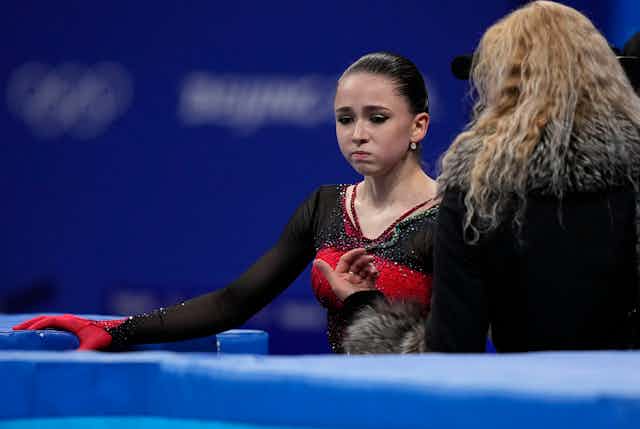 A skater in a black and red outfit with her hair pulled back and a woman with long wavy blond hair seen from behind