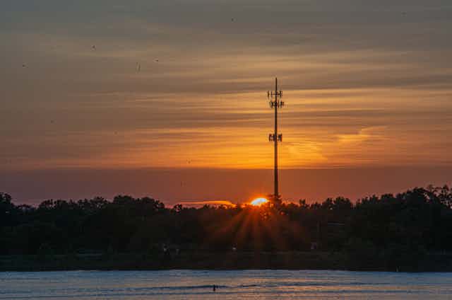A cell phone tower rises above a tree line with water in the foreground and the sun setting behind the trees near the tower
