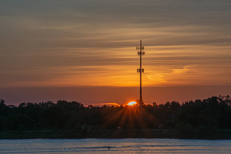 A cell phone tower rises above a tree line with water in the foreground and the sun setting behind the trees near the tower