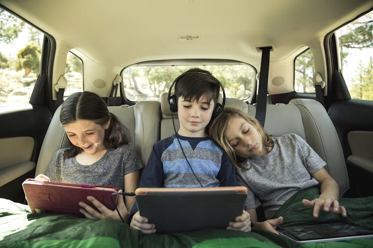 Three children in the backseat of a car use tablet devices