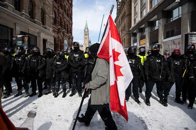 A man with a flag walks in front of a police line while the Peace Tower of the Parliament Buildings can be seen in the background.