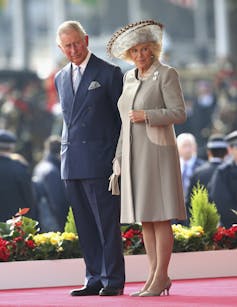A grey-haired man in a blue suit stands next to a woman in a beige coat and hat.