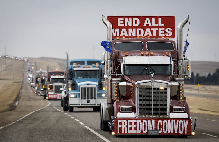 Several trucks are seen on a highway, with the first in line adorned with signs that say END ALL MANDATES and FREEDOM CONVOY