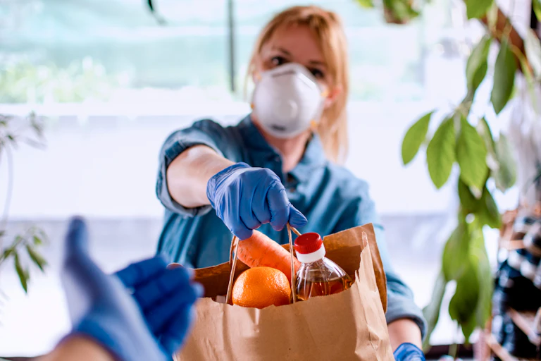 Une femme tend un sac de provisions alimentaires (carottes, oranges…) dans un sac en papier