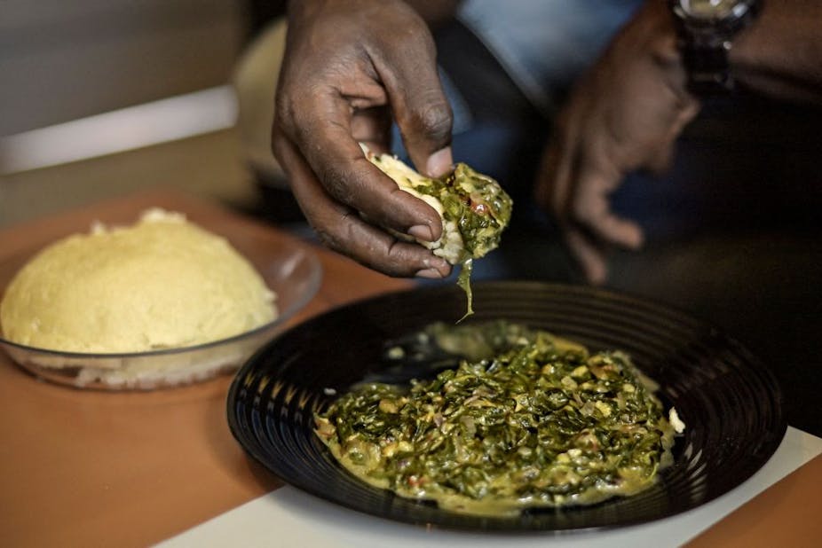 Two plates of food, one with a mound of porridge and the other a green stew. A man's hand scoops the two together.