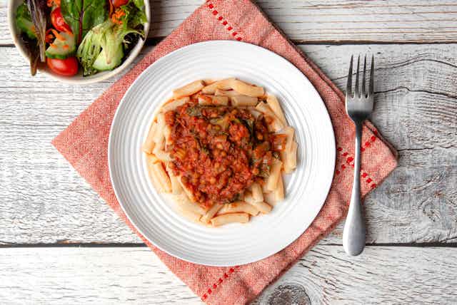 A pasta dish on a white plate, resting on a red napkin on a wooden table with a fork to the right and a salad top left