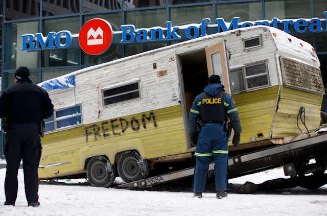 a camper with 'freedom' spraypainted on its side gets towed from a street in front of a bank of montreal
