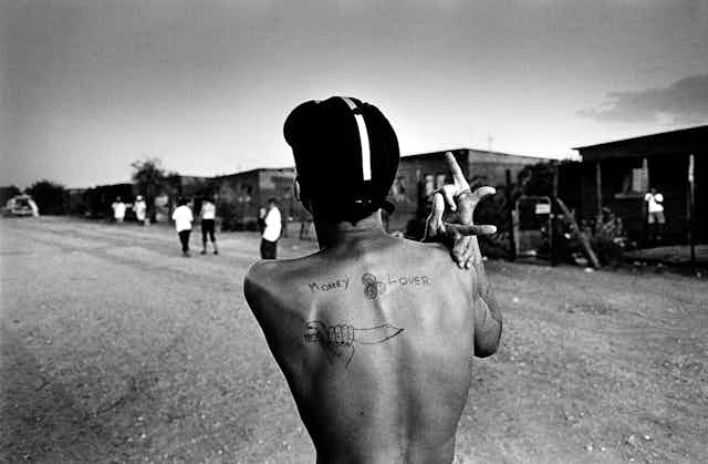 A black and white image of a young man from behind. He stands on a township street, a cap on his head, his raised fingers forming an intricate shape and a tattoo displayed on his back showing a knife and the words, 'Money Lover'.