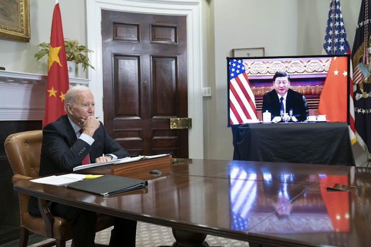 US president Joe Biden sits at a table in front of a Chinese flag, with Chinese president Xi Jinping on a video screen.