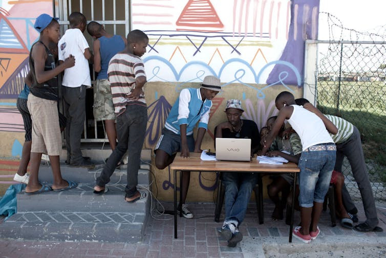 A group of boys gathers at a club house, one sitting and entering information into a computer at an outside table, others standing around him or peering through the security gate of the clubhouse, which sports painted murals of football players.